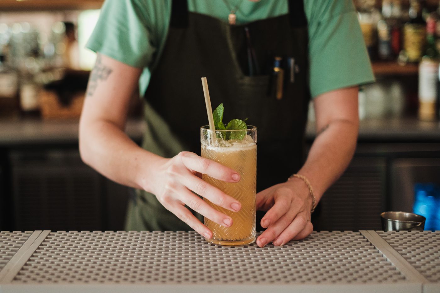 A bartender in an apron serving a long cocktail with mint across the bar